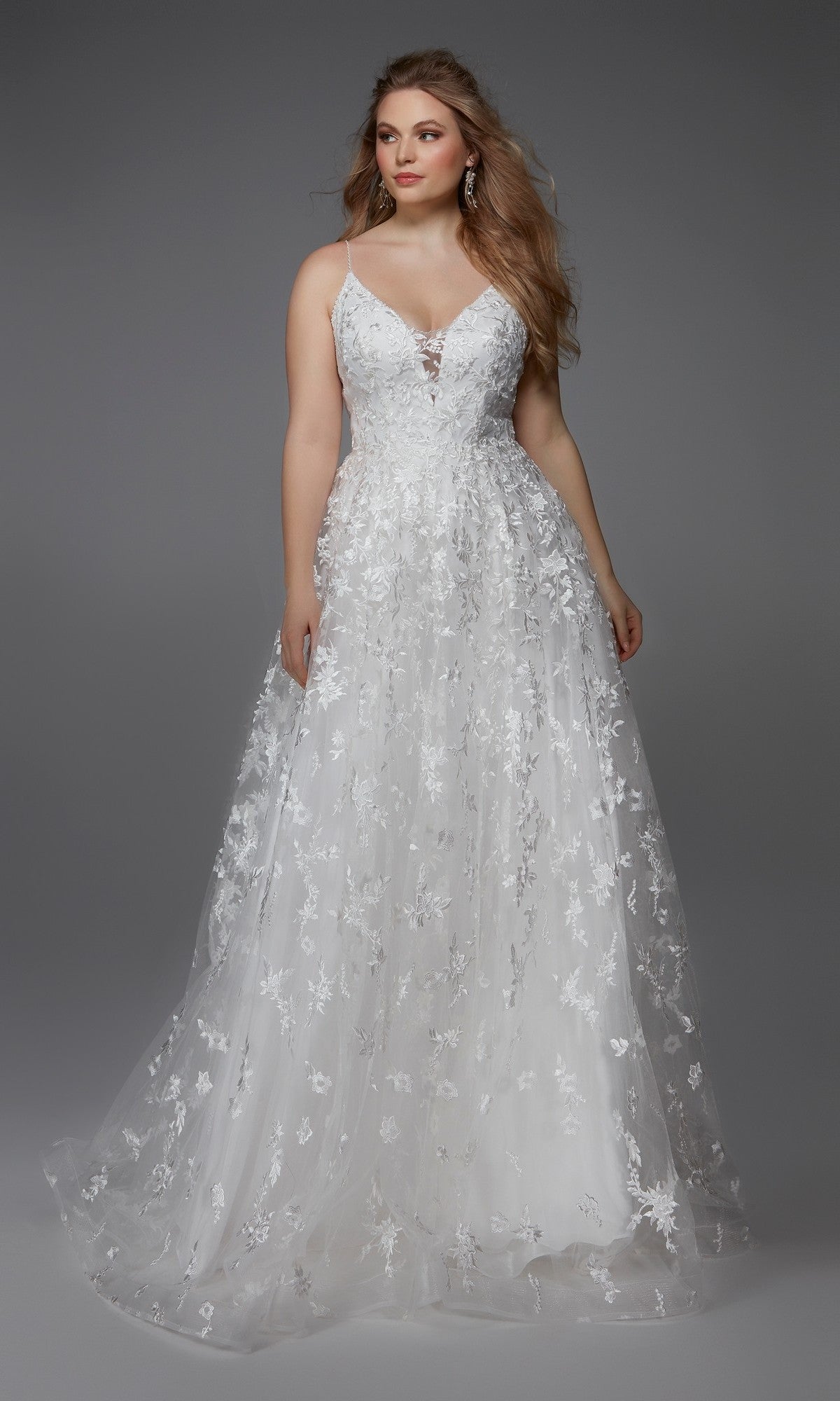 A woman stands against a plain gray background wearing the Alyce 7012 long formal dress, featuring an A-line silhouette, white floor-length design, fitted bodice, and floral lace appliques.